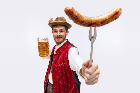 Joyful man wearing traditional fest Bavarian or German outfit with big beer glass showing fried sausage on fork. Oktoberfest, party, holiday, ad, sales conceptの写真素材