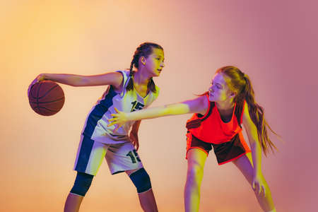 Attack and defense. Female basketball players, young girls, teen in action with basketball ball isolated on neoned background. Concept of sport, team, enegry, competition, skillsの写真素材