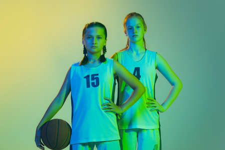 Two sportive girls, teens, basketball players standing together with basketball ball isolated on green background in neon light. Concept of sport, team, enegry, competition, skillsの写真素材