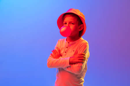 Studio shot of stylish little boy, kid posing isolated on gradient blue-purple background in neon light. Concept of child emotions, facial expression, fashion and ad.の写真素材