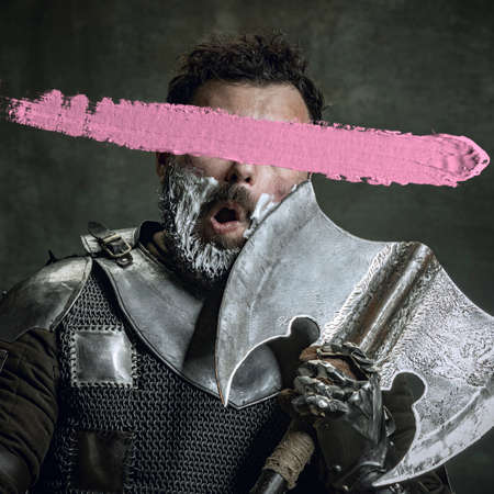 Artwork. Young man as medieval warrior, knight isolated on dark vintage background. Random stroke of pink paint on his face. Art, creativity, eras comparison concept.の写真素材