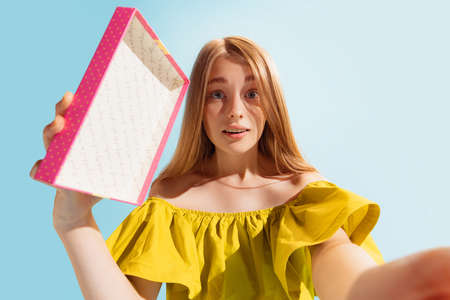 Wide angle bottom view of young beautiful astonished girl holding gift box isolated on blue background. Concept of emotions, holidays, inspiration, sales, ad.の写真素材