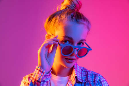 Pretty young girl, student in cool eyeglasses wearing plaid shirt isolated on magenta color background in neon light. Concept of beauty, art, fashion, emotionsの写真素材