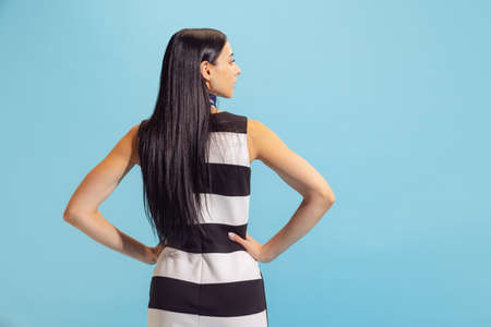 Back view of adorable young woman in summer striped dress posing isolated on blue background. Concept of beauty, art, fashion, human emotionsの写真素材