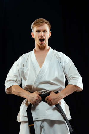 Portrait of young man, karate fighter in white kimono and black belt in fighting stance on dark background. Sport, action, combat sports, ad conceptの写真素材