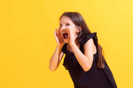 Shouting little girl, kid wearing black dress shouting isolated on yellow background. Concept of children emotions, fashion, beauty, school and adの写真素材
