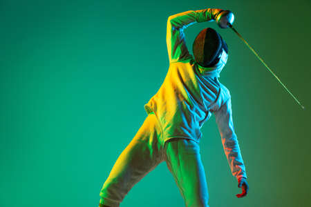Studio shot of young man, fencer with smallsword practicing fencing isolated on green background in neon light. Sport, energy, skills, achievementsの写真素材