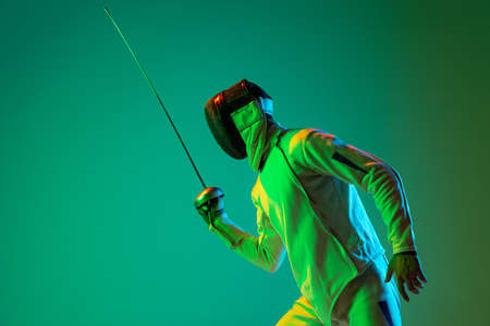 Studio shot of young man, fencer with smallsword practicing fencing isolated on green background in...