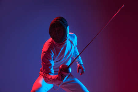 Male fencer with smallsword practicing fencing isolated on purple background in neon light. Sport,...