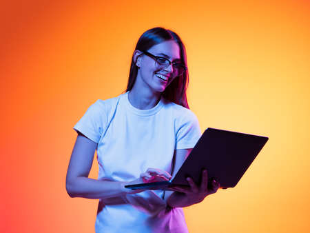 Portrait of young smiling girl, student in white t-shirt with laptop isolated on orange color background in neon light. Concept of beauty, fashion, emotionsの写真素材
