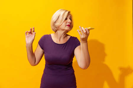 Emotional senior woman in purple dress using cellphone isolated on bright yellow background. Concept of beauty, age, emotions, modern gadgetsの写真素材
