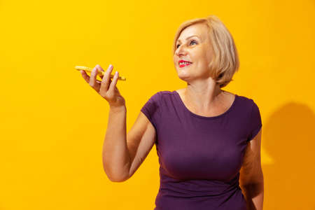 Emotional senior woman in purple dress using cellphone isolated on bright yellow background. Concept...