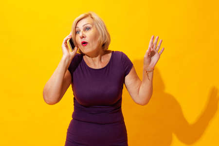 Emotional senior woman in purple dress using cellphone isolated on bright yellow background. Concept of beauty, age, emotions, modern gadgetsの写真素材