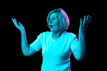 Surprised senior woman with blond hair raising hands up in surprise isolated over dark background in...