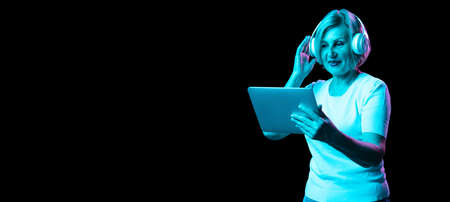 Emotional senior woman in white t-shirt using digital tablet isolated on dark background in neon light. Concept of beauty, age, emotions, modern gadgetsの写真素材