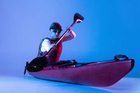 Water sport activities. Sportive woman in red canoe, kayak with a life vest and a paddle isolated on blue background in neon light. Concept of sportの写真素材