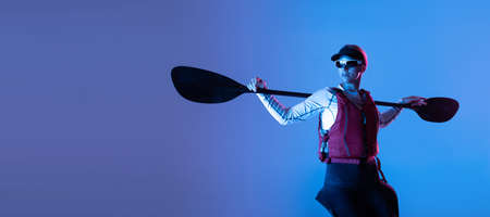 Water sport activities. Sportive woman in red canoe, kayak with a life vest and a paddle isolated on blue background in neon light. Concept of sportの写真素材
