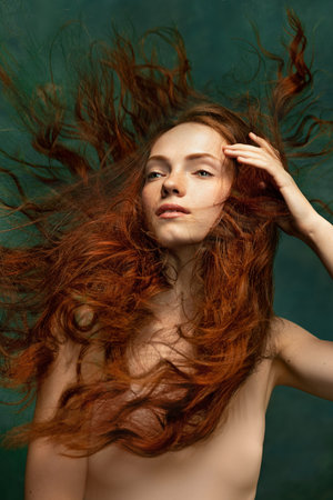 Young attractive redhead girl with long curly hair wearing nude color lingerie isolated over dark...