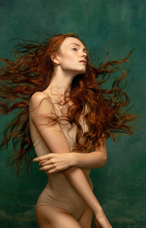 Young attractive redhead girl with long curly hair wearing nude color lingerie isolated over dark green background. Magic look. Concept of beauty, artの写真素材