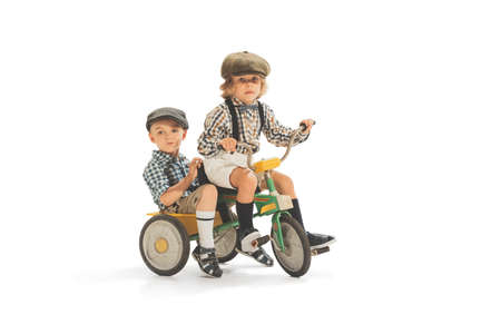 Cute happy kids riding tricycle isolated on white studio background. Retro vintage style concept. Friendship, hobbies, art, eras comparisonの写真素材