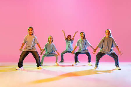 Portrait of cheerful, active little girls, happy kids dancing isolated on orange background in neon light. Concept of music, fashion, art, childhood, hobbyの写真素材