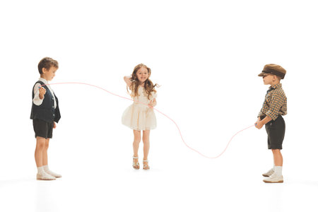 Portrait of three children, boys and girl in retro clothes playing together, jumping over rope isolated over white studio backgroundの写真素材