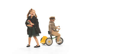 Portrait of cheerful girl with books playing with little boy on small bike isolated over white...