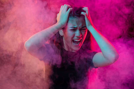 Emotional pain. Young dark hair woman shouting isolated over neoned pink background with clouds of smoke. Concept of mental health, art, emotionsの写真素材