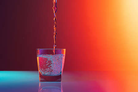 Composition with water flows into glass placed on table over gradient multicolored background in...