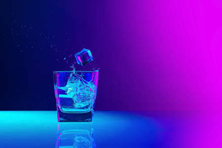Ice cube falls into a transparent glass of water standing on mirror surface over gradient blue-pink background in neon light. Art, beauty, drinksの写真素材