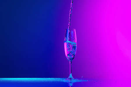 Authentic hand pouring pure water splash from wine or champagne glass over bright blue-pink gradient background in neon light. Art, drinks, creativityの写真素材