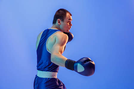 Practicing punch. Muscled man, male boxer in blue uniform and boxing gloves training isolated on navy background in neon.の写真素材