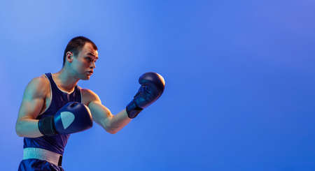 Muscled man, male boxer in blue uniform and boxing gloves training isolated on navy background in...
