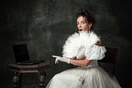 Shock. Creative portrait of young beautiful girl elegant dress of medieval fashion style with veer isolated on dark background. Comparison of eras concept, flemish style.の写真素材