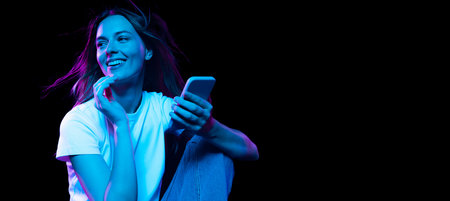Happy young pretty girl with cellphone isolated on dark background in purple neon light. Concept of...
