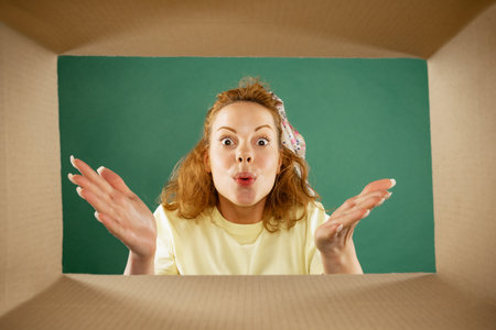 Beautiful young girl opening, unpacking carton box and looking inside with surprise. Emotions, black friday, holidays, online shopping conceptの写真素材