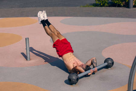 Young strong muscular man doing strength exercises on workout ground at public park in summer...