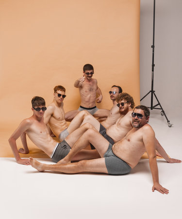Team of young handsome men of different body types sitting together in underwear on floor at studio photo shoot. Fun, joy, body positive and emotionsの写真素材