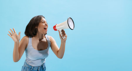 Excited young girl, student wearing casual style clothes shouting at megaphone isolated on light blue background in neon light.の写真素材