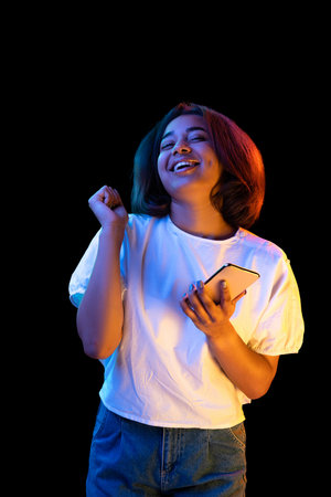 One astonished glad woman in white t-shirt using smart phone with amazed expression isolated on dark background in neon.の写真素材