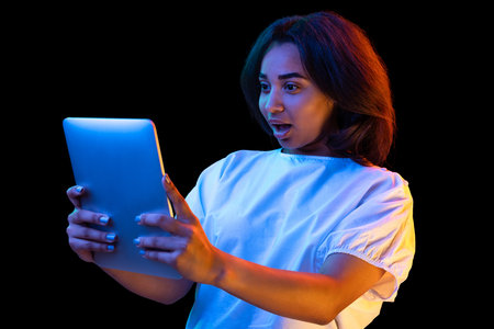 Portrait of young shocked girl, student in white t-shirt with digital tablet isolated on dark background in neon light. Concept of studying, fashion, emotionsの写真素材