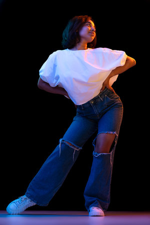 Young charming african girl, student in white t-shirt and jeans posing isolated on dark background in neon. Concept of beauty, fashion, emotions, youthの写真素材