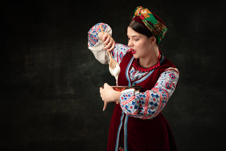 Emotional young woman wearing national folk Ukrainian attire isolated over dark retro background. Emotions, fashion, beauty, eras comparison conceptの写真素材