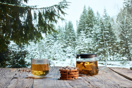 Glass pot with herbal tea, cakes, biscuits on wooden table over winter snow forest background. Design for greeting card. Holidays, vacation, traditionsの写真素材