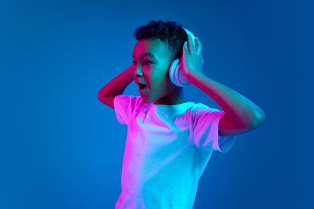 Little cheerful happy boy 6-7 years old wearing white t-shirt and headphones listen to music isolated on blue background in neon light.の写真素材