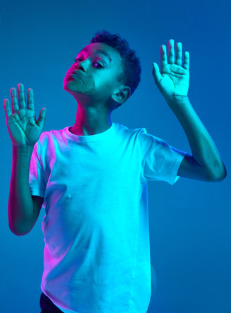 Close-up of little boy leaning against transparent glass by cheek isolated on blue background in neon light. Funny face. Concept of emotionsの写真素材