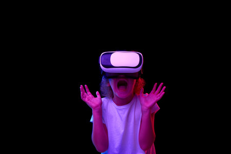 School age girl in white t-shirt enjoying 3D technology in virtual reality headset isolated on dark...