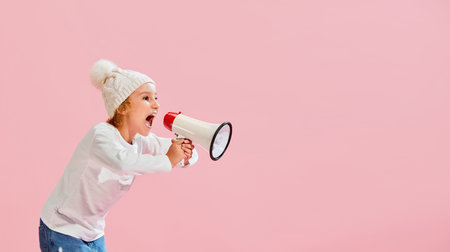 Studio shot of little girl, kid in casual style clothes and warm hat shouting at megaphone isolated over pink background.の写真素材