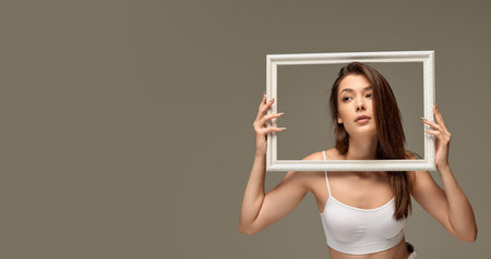 Portrait of young beautiful woman posing, looking into picture frame isolated over grey studio backgroundの写真素材