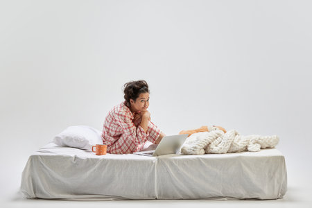 Portrait of young charming girl in home wear sitting on bed at home isolated on gray background. Ideas, inspiration, imagination. Art, beauty, youthの写真素材